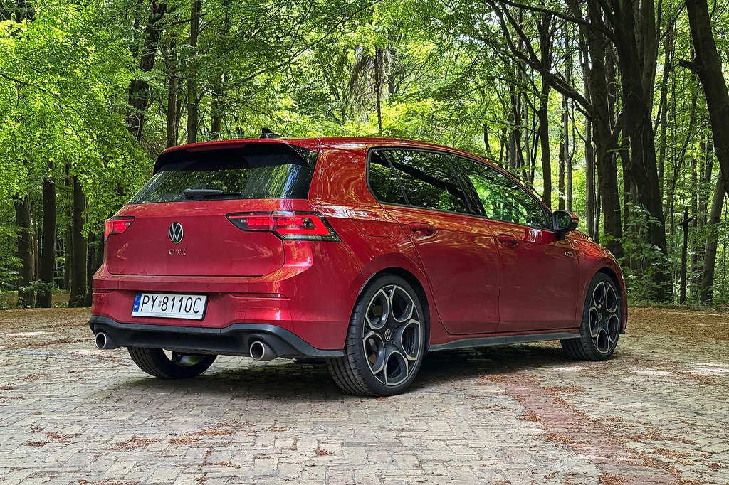 Katalogowa cena nowego Golfa GTI wynosi 167 890 zł. Sporo, ale taka jest cena dobrej zabawy. Czerwony samochód Volkswagen Golf GTI zaparkowany na wybrukowanym placu w otoczeniu zielonego, gęstego lasu, widok od tyłu lekko z boku.
