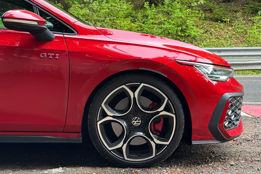 19-calowe felgi Queenstown idealnie współgrają z linią nadwozia sportowego Golfa. Czerwony samochód Volkswagen GTI zaparkowany przy barierce ochronnej, eksponując przednie koło, felgę o nowoczesnym wzorze i charakterystyczne logo marki