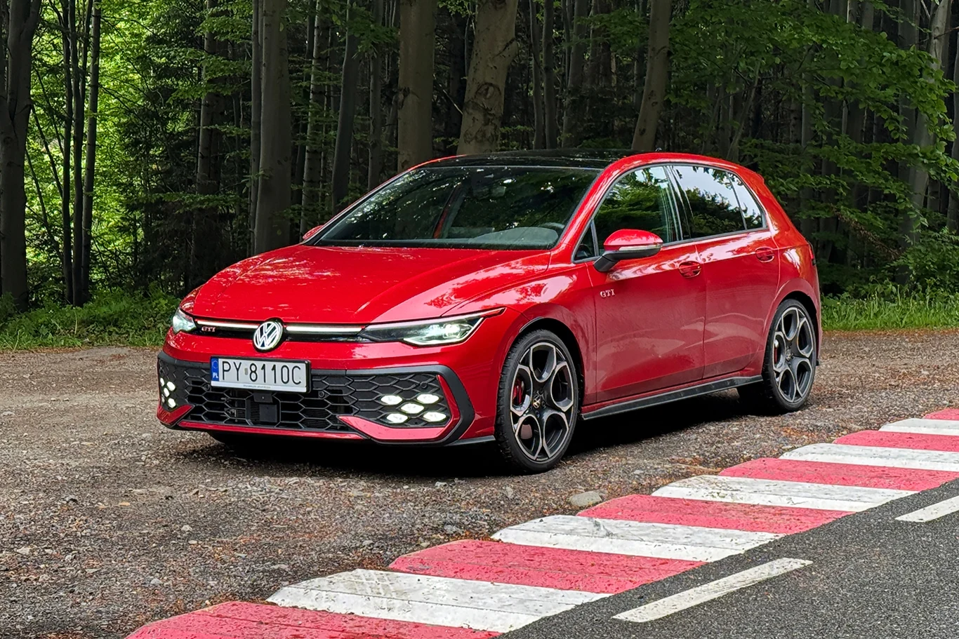 Nowy Golf GTI przyspiesza szybciej od poprzednika. 0 do 100 km/h "robi" teraz w 5,9 sekundy. Czerwony samochód osobowy marki Volkswagen zaparkowany przy krawędzi asfaltowej drogi o wyścigowym malowaniu, w tle gęsty las.