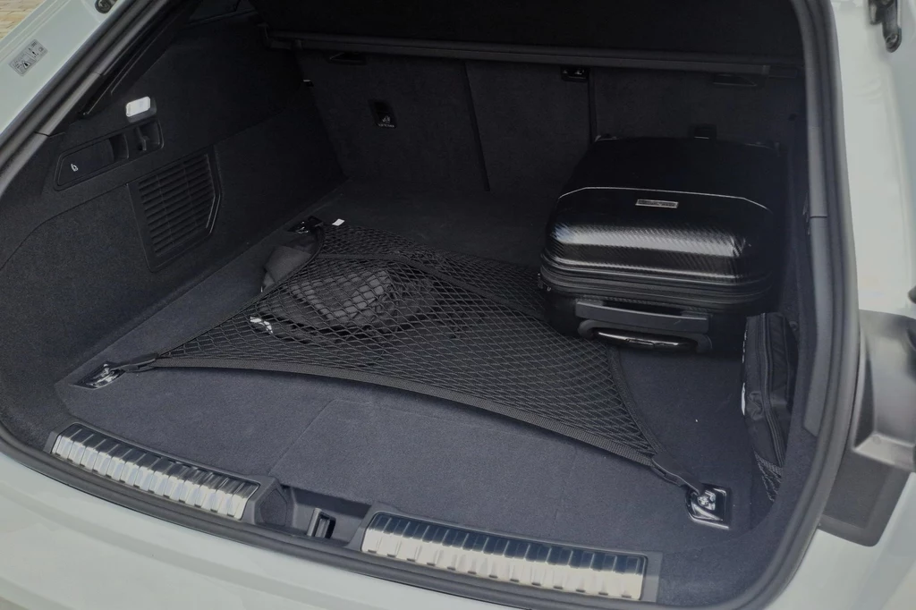 Bagażnik w Audi A5 e-hybrid, z uwagi na obecność akumulatora, jest mniejszy niż w pozostałych wariantach napędowych modelu. Bagażnik samochodu z czarną walizką umieszczoną pod siatką zabezpieczającą, wnętrze czyste i dobrze uporządkowane.