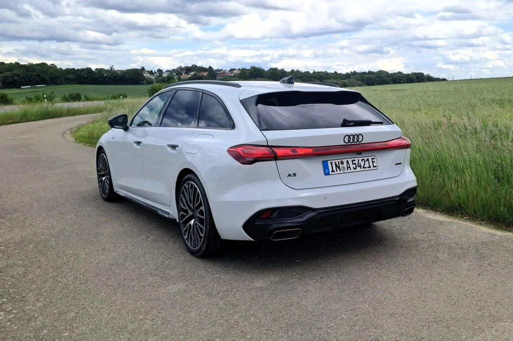 Audi A5 w trybie "hybrid" może doładowywać akumulator za pomocą silnika spalinowego tylko powyżej prędkości 65 km/h. Białe Audi A5 w wersji kombi zaparkowane na bocznej, asfaltowej drodze, otoczonej zielonymi polami i niską roślinnością, krajobraz wiejski w tle z lekko pochmurnym niebem.