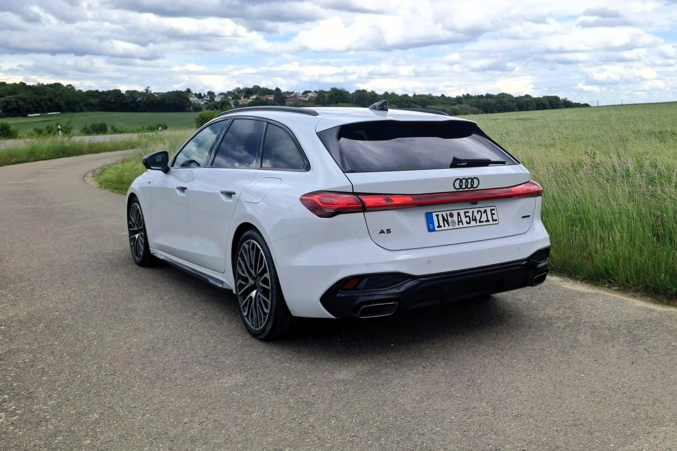 Białe Audi A5 w wersji kombi zaparkowane na bocznej, asfaltowej drodze, otoczonej zielonymi polami i niską roślinnością, krajobraz wiejski w tle z lekko pochmurnym niebem.