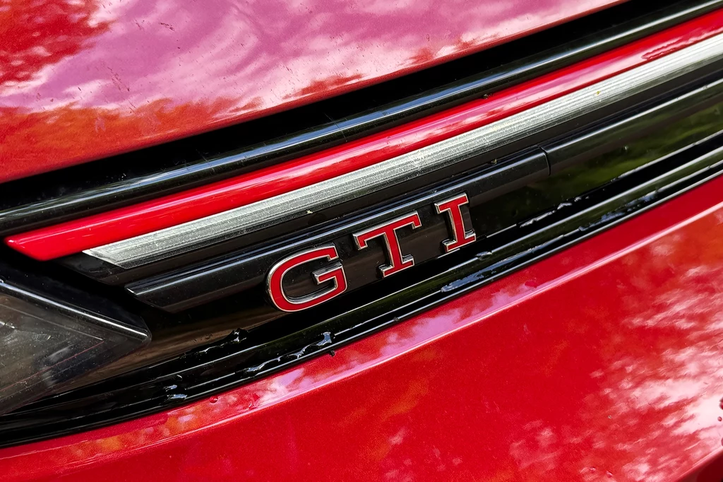 Zaprezentowany w 1976 roku Golf GTI, szybko stał się punktem odniesienia dla wielu przyszłych hot hatchy. Czerwony grill samochodu z widocznym emblematem GTI na czarnym pasku, detale nadwozia podkreślone refleksami światła.