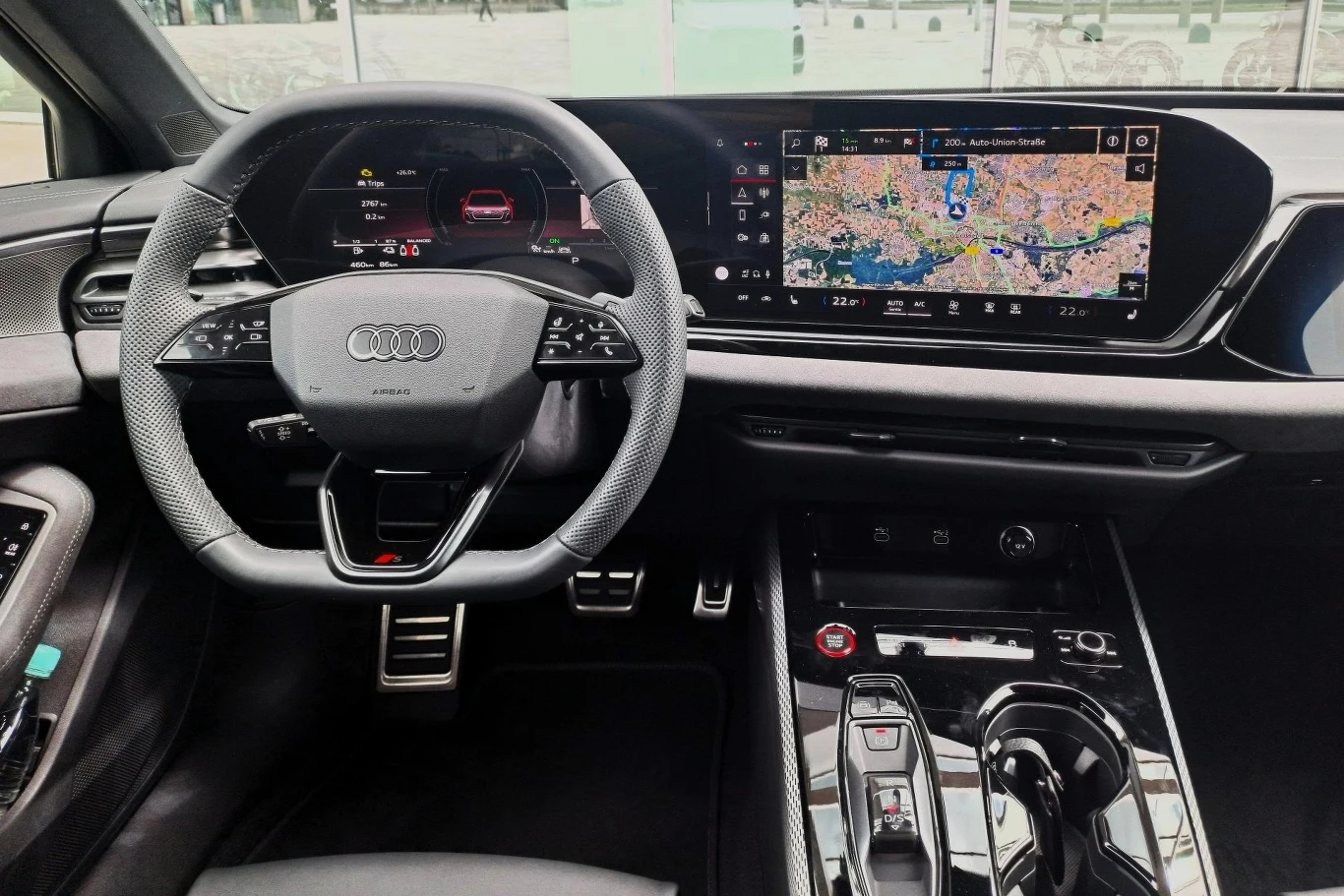 Luksusowe wnętrze samochodu marki Audi z widocznym nowoczesnym kierownicą, cyfrowym panelem wskaźników oraz dużym ekranem systemu multimedialnego z nawigacją GPS pokazującą mapę miasta.