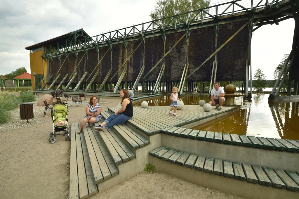 Drewniane schody i podesty nad stawem z wodą, kilka osób siedzi i odpoczywa, w tle duża konstrukcja przypominająca tężnię solankową, obok wózek dziecięcy, z tyłu drzewa i zabudowania.