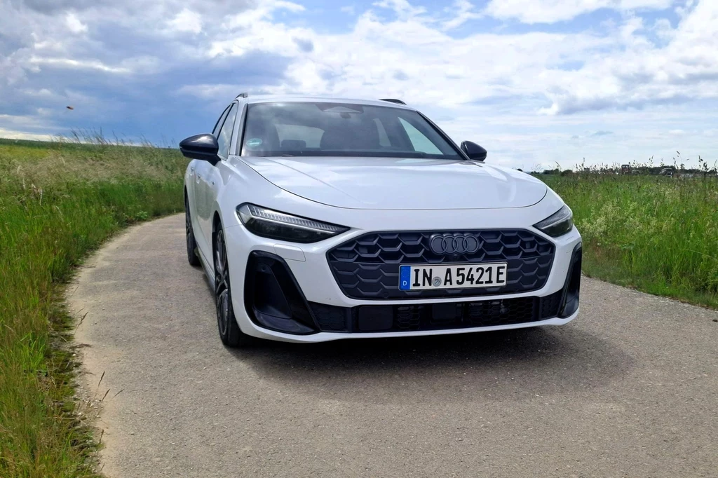 Audi A5 z hybrydą plug-in dostępne jest w dwóch wariantach mocy - 299 KM i 367 KM. W obu napęd trafia na cztery koła. Biały samochód marki Audi stoi na wąskiej asfaltowej drodze, otoczonej zielonymi łąkami, pod lekko zachmurzonym niebem.