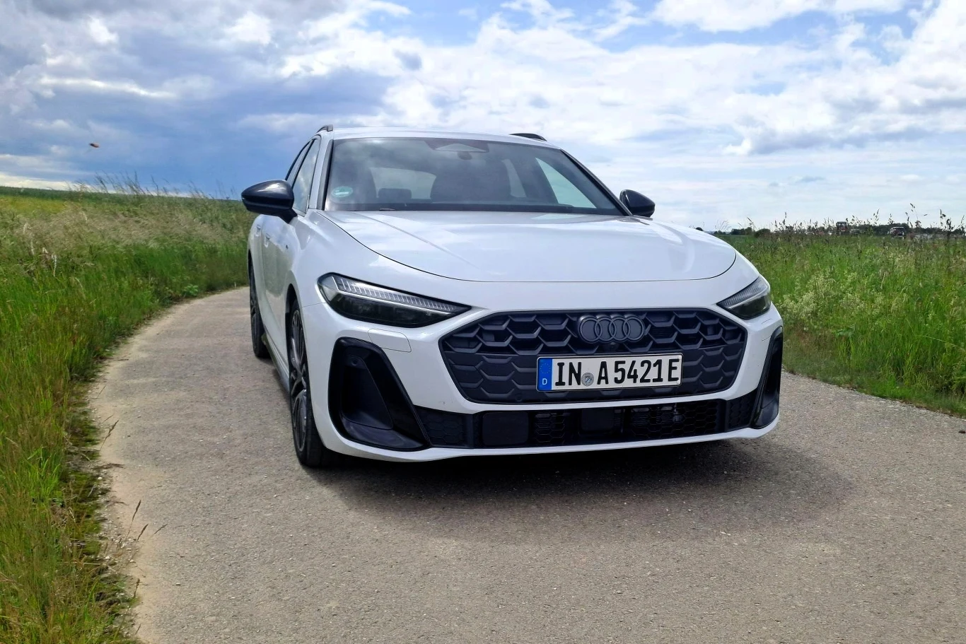 Biały samochód marki Audi stoi na wąskiej asfaltowej drodze, otoczonej zielonymi łąkami, pod lekko zachmurzonym niebem.
