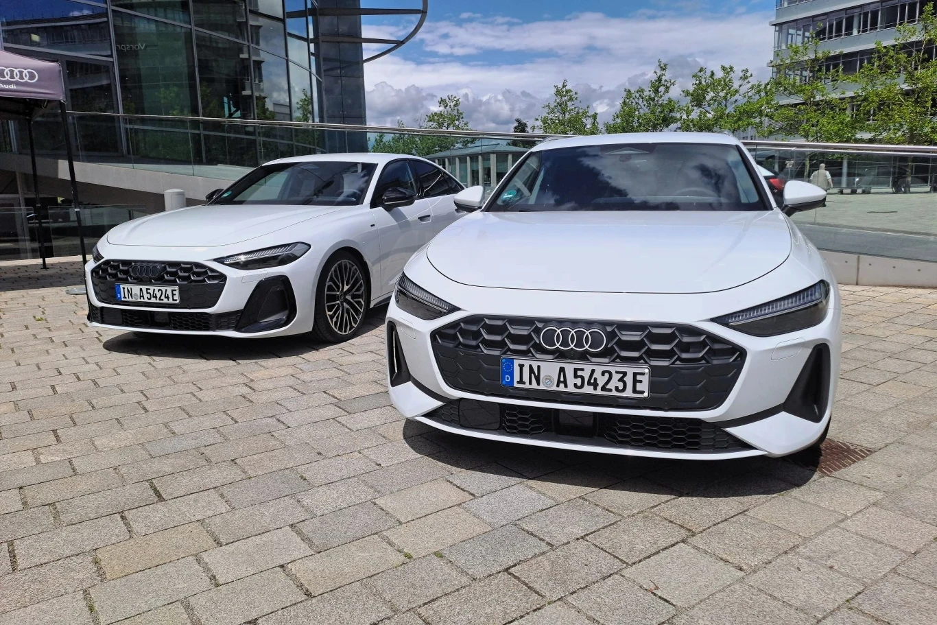 Dwa nowoczesne samochody marki Audi w białym kolorze zaparkowane na wybrukowanym placu przed nowoczesnym budynkiem o szklanych fasadach, z widocznymi drzewami i elementami miejskiej architektury w tle.