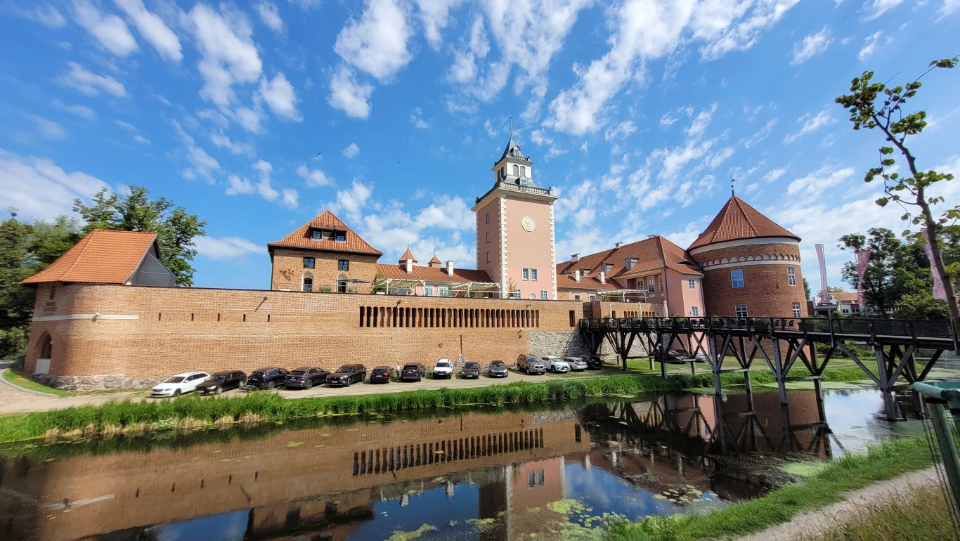 Imponujący Zamek Biskupów Warmińskich w Lidzbarku Warmińskim Ceglany zamek z wieżą i charakterystycznymi dachami, otoczony murami obronnymi oraz fosą odbijającą budowlę, przed zamkiem parkowane samochody, niebo z drobnymi chmurami.