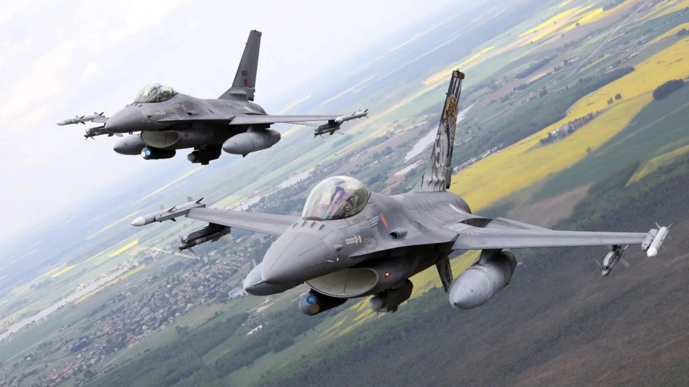 Tajemniczy atak Rosji. Grad pocisków spadł na bazę myśliwców F-16? Dwa myśliwce F-16 lecą w formacji nad polami i lasami, w tle widoczny krajobraz wiejski z polami uprawnymi i zabudowaniami.
