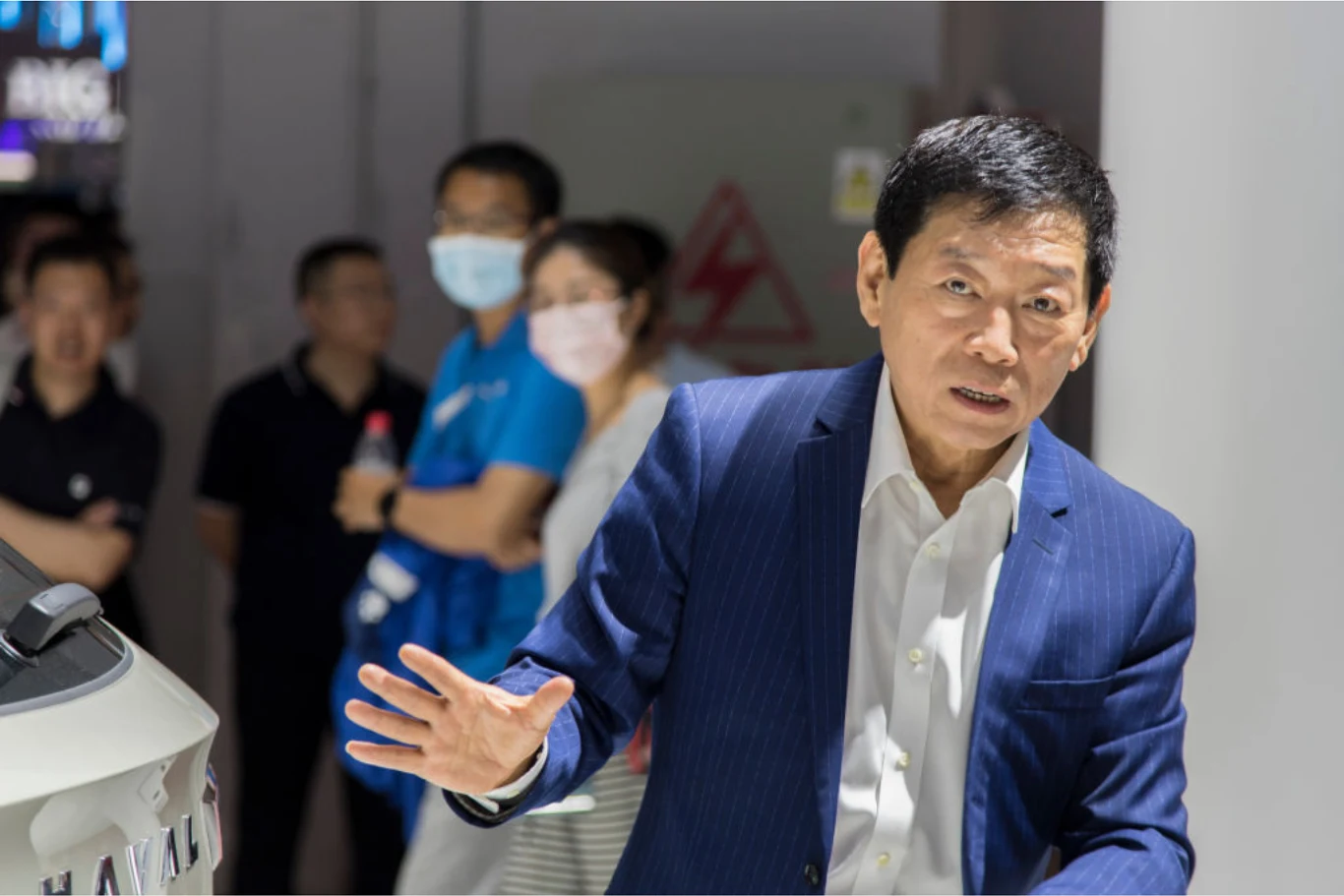 Mężczyzna w garniturze z wyciągniętą ręką, w tle kilka osób, część z nich w maseczkach, a także biały obiekt z napisem HAVAL.