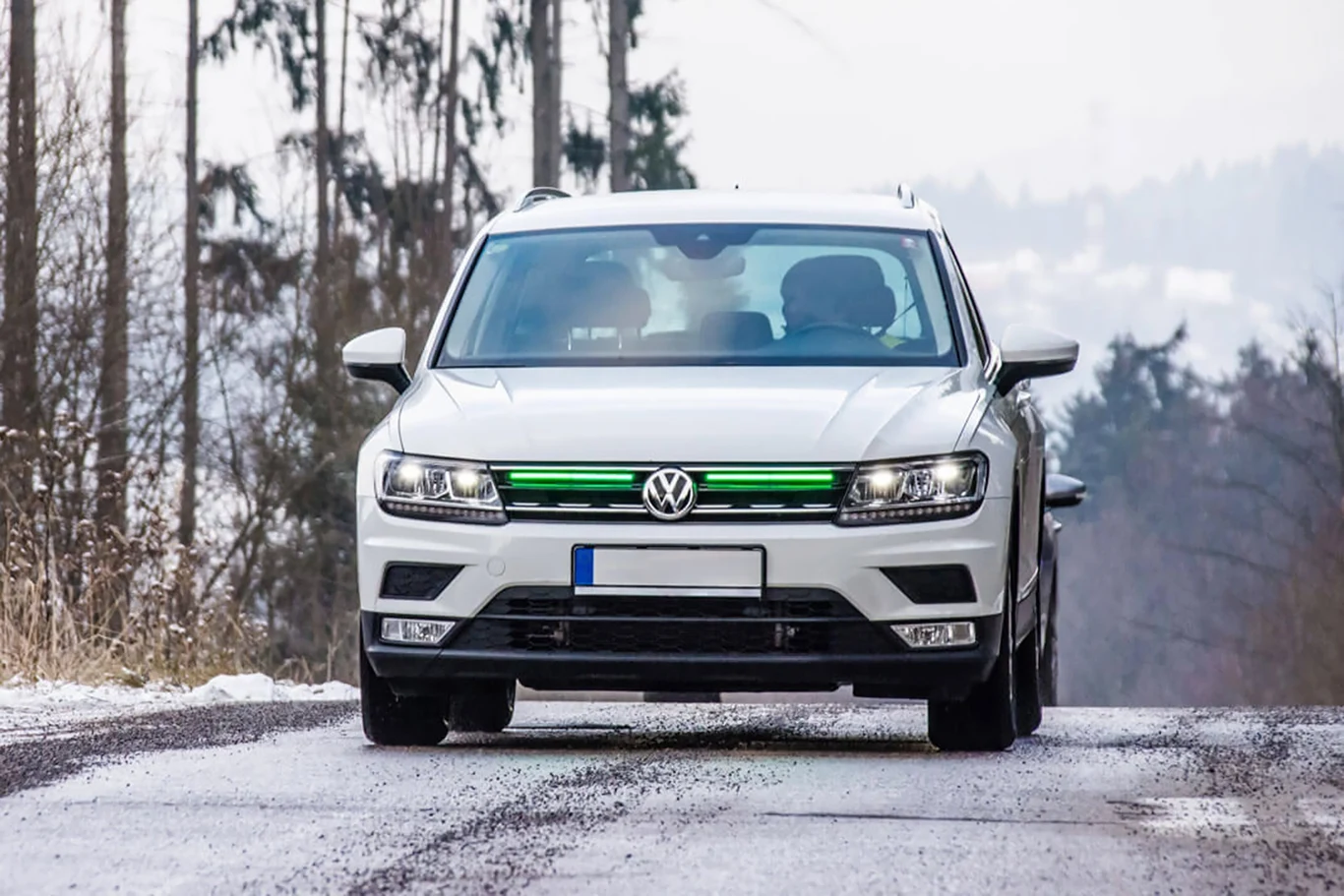 Zielone, przednie światło stopu. Nowy typ oświetlenia w autach | Fot. frontbrakelights.com Biały samochód osobowy marki Volkswagen jedzie po ośnieżonej, leśnej drodze, wokół znajdują się drzewa, a w tle widoczna jest lekko zamglona panorama.