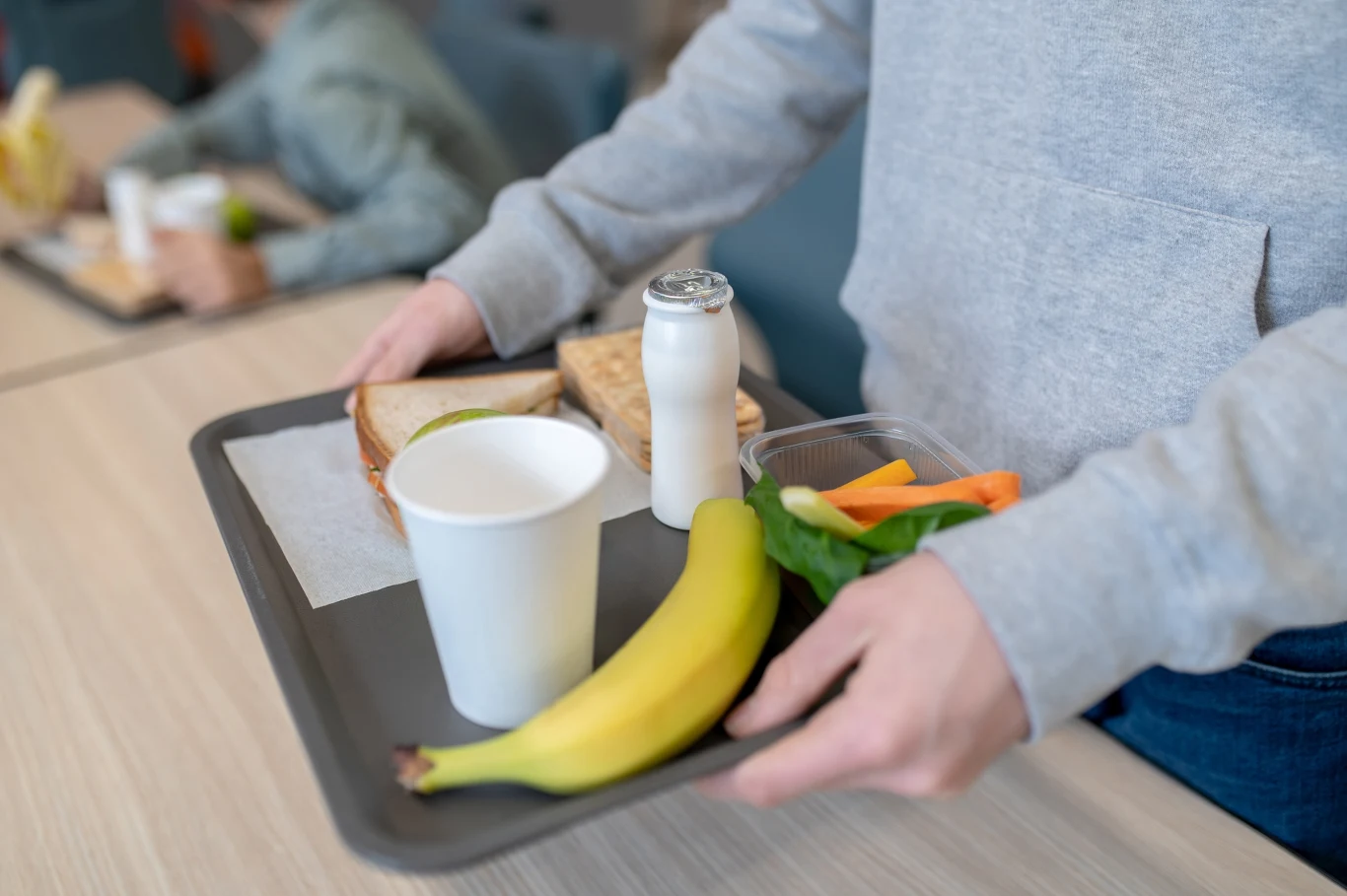 Taca z przygotowanym posiłkiem zawierającym banana, białą tekturową filiżankę, butelkę napoju mlecznego, kanapkę oraz pudełko z pokrojonymi warzywami; osoba ubrana w szarą bluzę trzyma tacę na jasnym stole.