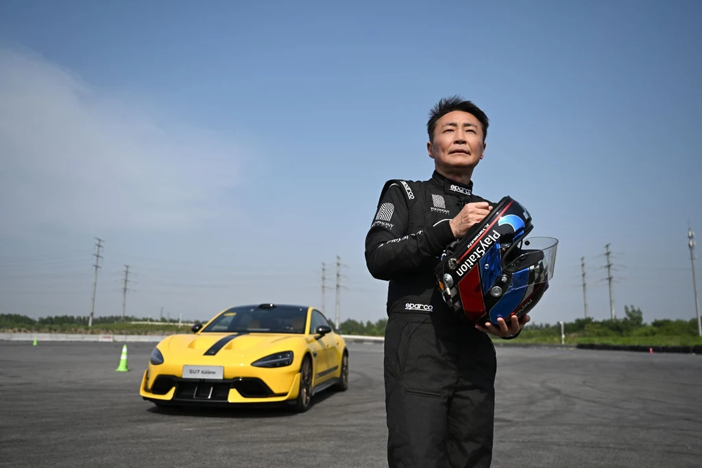 Kazunori Yamauchi przetestował SU7 Ultra i zdecydował. Mężczyzna w kombinezonie rajdowym trzymający kask wyścigowy na tle żółtego sportowego samochodu zaparkowanego na placu o asfaltowej nawierzchni, w oddali widać niskie krzewy i linie wysokiego napięcia.