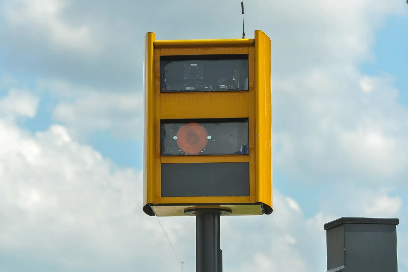 Żółty fotoradar zamontowany na wysokim słupie na tle nieba pokrytego chmurami.