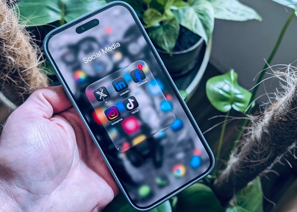 Apple Liquid Glass na iPhonie. Nowy design trafi też na inne urządzenia Apple'a Nowoczesny smartfon z widocznym ekranem prezentującym folder z ikonami popularnych aplikacji społecznościowych takich jak X, LinkedIn, Facebook, Instagram, Messenger oraz TikTok, trzymany w dłoni na tle zielonych roślin doniczkowych.