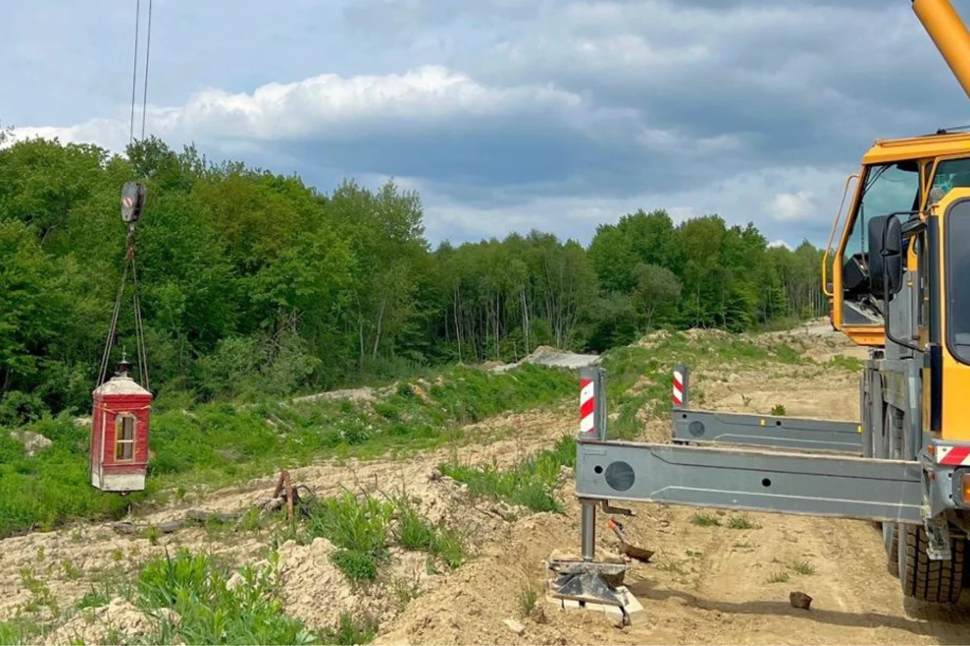 Drogowcy musieli przenieść zabytkową kapliczkę. Dźwig podnosi małą, czerwoną budkę telefoniczną w otoczeniu zieleni i nieużytków, na tle lasu i nieba z chmurami, po prawej widoczna część pojazdu dźwigu.