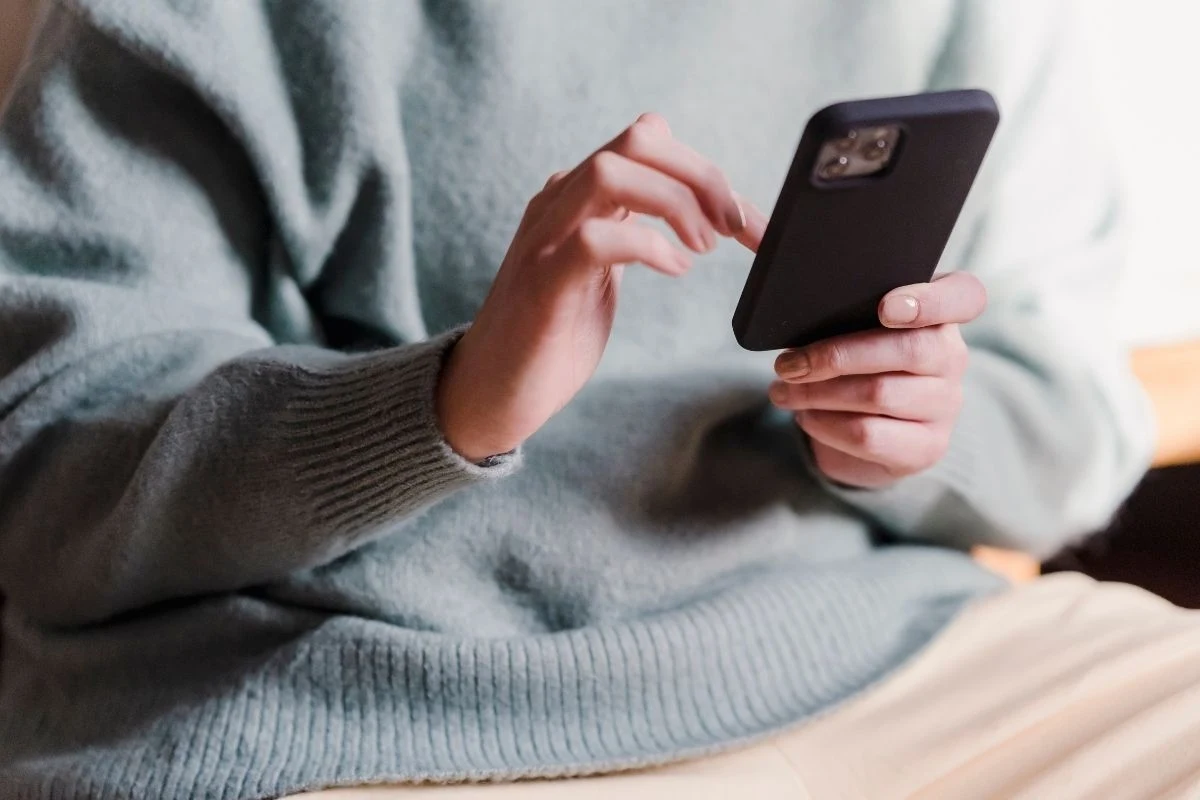 TikTokowe filmiki z poradami dot. zdrowia psychicznego szkodzą i dezinformują Osoba w zielonym swetrze trzyma smartfon w obu dłoniach i korzysta z ekranu dotykowego, siedząc w jasnym pomieszczeniu.