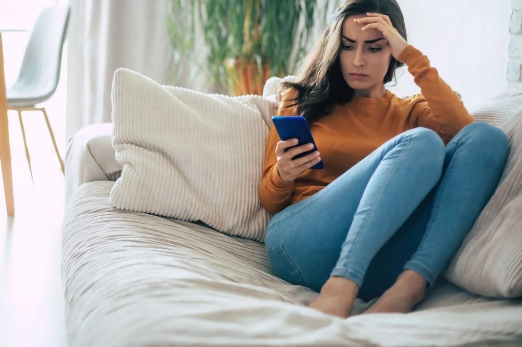 Filmiki internetowych twórców lepiej omijać albo brać na nie solidną poprawkę Młoda kobieta siedząca na sofie, ubrana w pomarańczowy sweter i niebieskie dżinsy, z zaniepokojonym wyrazem twarzy patrzy na ekran smartfona, trzymając jedną rękę na czole.
