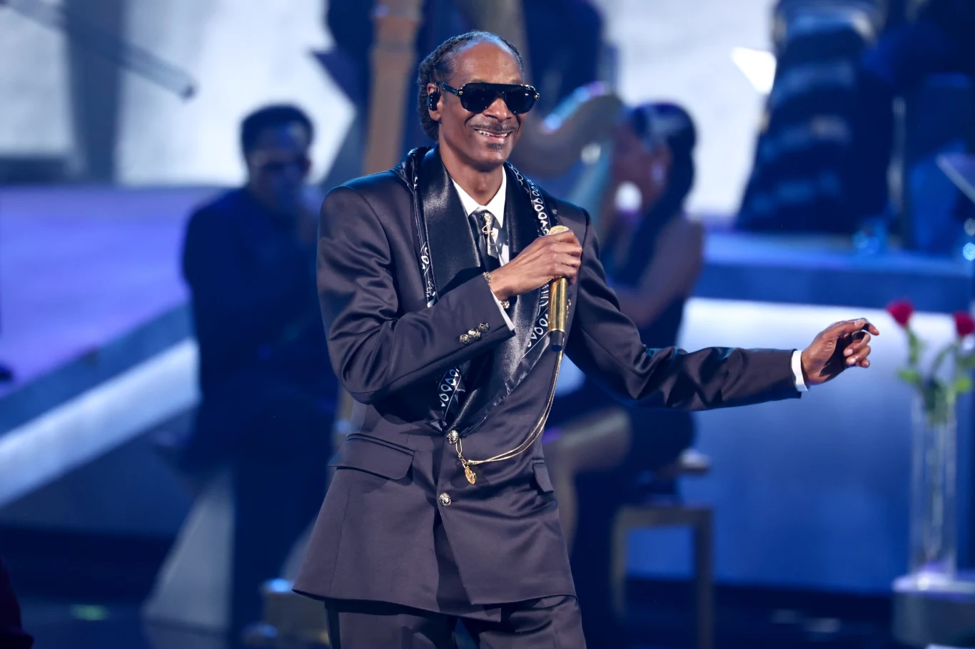 Snoop Dogg podczas występu na BET Awards 2025 Mężczyzna w ciemnych okularach i eleganckim garniturze śpiewa lub przemawia do mikrofonu na scenie koncertowej przy nastrojowym oświetleniu, w tle widoczny rozmyty zarys publiczności i instrumentów.
