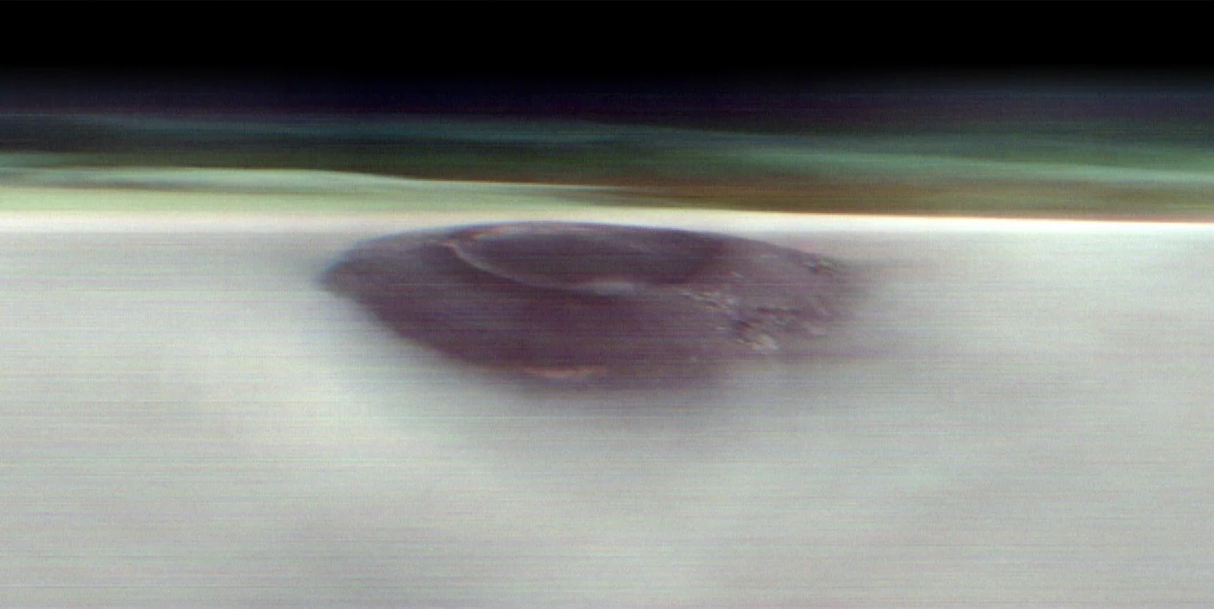 Arsia Mons to jeden z najwyższych wulkanów na Marsie Rozmyty, ciemny obiekt o okrągłym kształcie znajduje się na jasnej, płaskiej powierzchni, w tle widoczne poziome pasy w odcieniach zieleni i czerni, co nadaje zdjęciu abstrakcyjny charakter.
