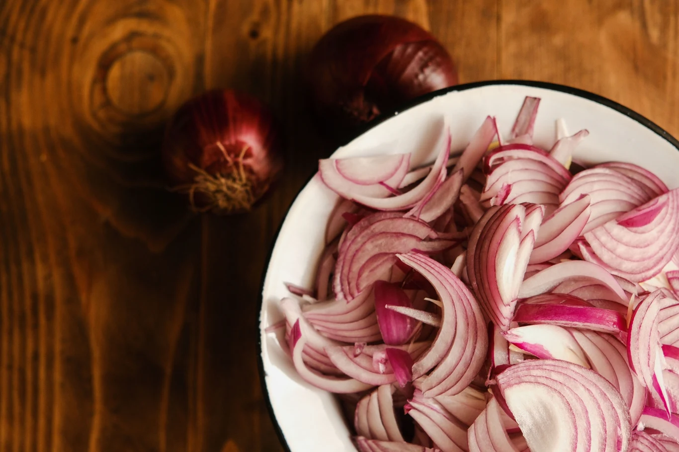 miska wypełniona pokrojoną w piórka czerwoną cebulą na drewnianym blacie, obok znajdują się dwie całe czerwone cebule