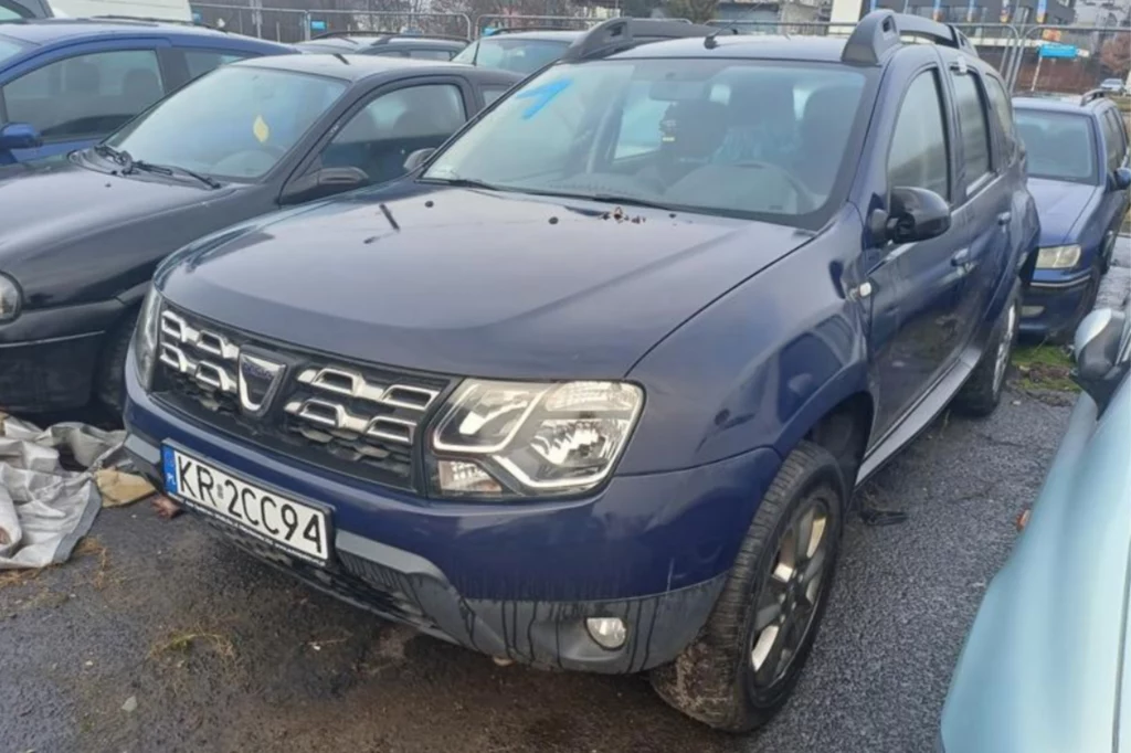 Granatowy Dacia Duster zaparkowany na parkingu, widoczny z przodu pod kątem. Samochód ma czarne felgi i srebrny grill. Na parkingu znajdują się inne samochody, a na ziemi leżą jakieś śmieci. Numer rejestracyjny samochodu to KR 2CC94