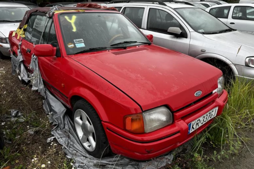 Czerwony Ford Escort Cabrio z otwartym dachem, częściowo pokryty folią ochronną. Samochód stoi na parkingu, prawdopodobnie na złomowisku, obok innych pojazdów. Na przedniej szybie widoczna jest żółta litera Y. Samochód ma polskie tablice rejestracyjne