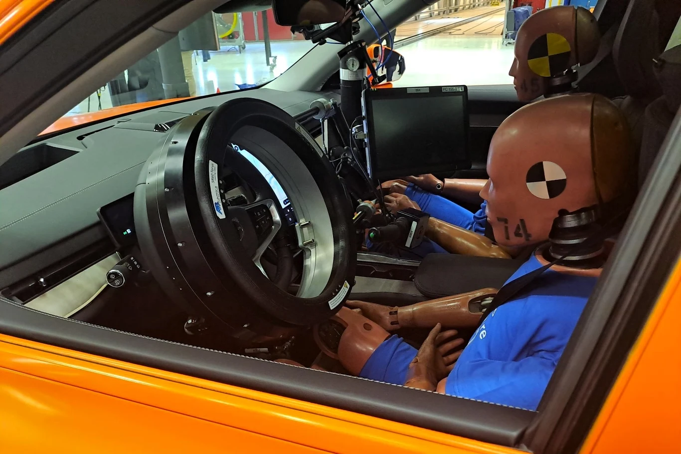 Dwa manekiny siedzą w samochodzie podczas testów zderzeniowych. Kierownica jest zautomatyzowana, a manekiny mają na sobie niebieskie koszulki i przypięte pasy bezpieczeństwa. Samochód jest pomarańczowy. Widać monitor wewnątrz samochodu. Można zauważyć ...