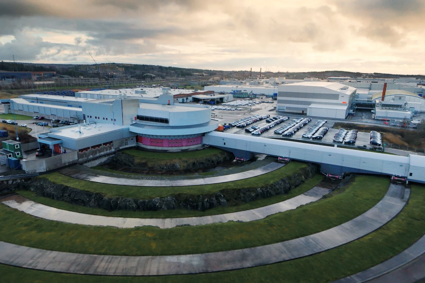 Widok z lotu ptaka na nowoczesny kompleks przemysłowy z parkingiem pełnym samochodów i otaczającą go zielenią. Kompleks składa się z kilku budynków o różnej architekturze, w tym okrągłego budynku z różowymi elementami. Na terenie kompleksu widoczne są ...