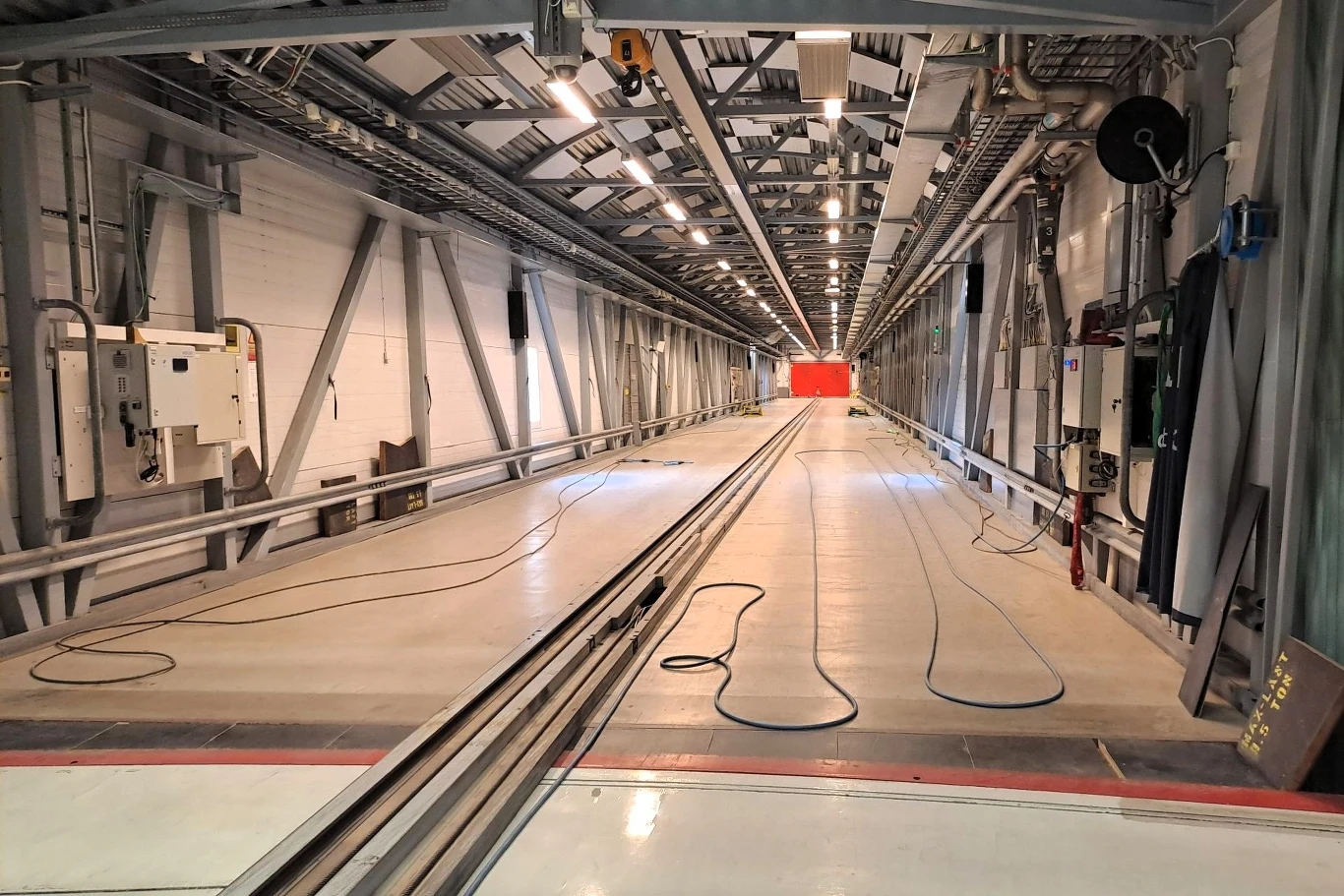 Długi, pusty korytarz z metalową konstrukcją i kablami na podłodze