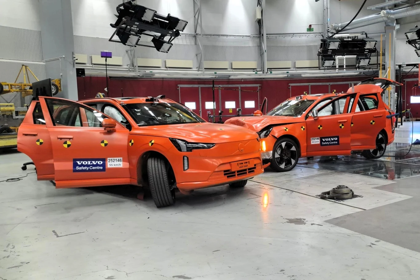 Dwa pomarańczowe samochody Volvo po teście zderzeniowym w centrum bezpieczeństwa Volvo. Jeden samochód ma otwarte drzwi i widoczne są uszkodzenia. Drugi samochód jest ustawiony pod kątem, a jego tył jest uszkodzony. W tle widać sprzęt do testów zderzen...