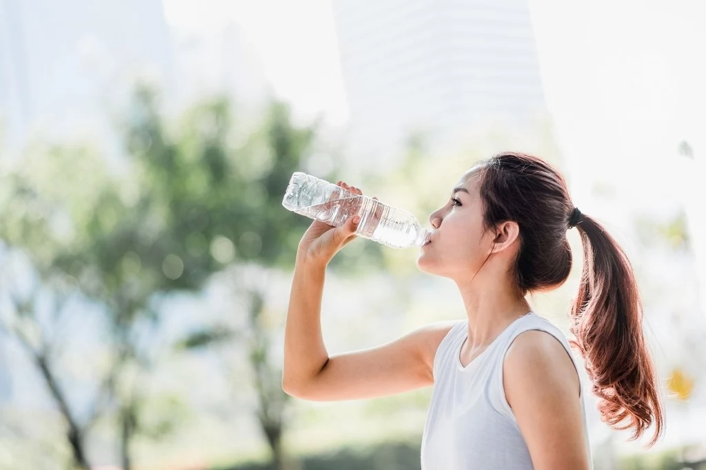 Niewystarczające nawadnianie się wpływa na nasz poziom energii w ciągu dnia Młoda Azjatka w sportowym stroju pije wodę z plastikowej butelki na tle zieleni miejskiego parku.
