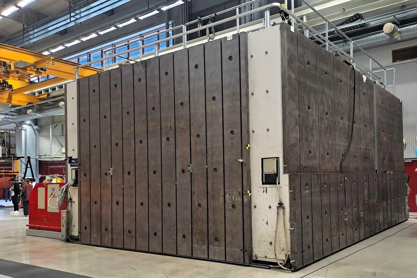 Duża, metalowa konstrukcja, przypominająca kontener lub komorę, stoi na betonowej posadzce w hali przemysłowej. Jej powierzchnia jest zbudowana z pionowych, metalowych płyt, z widocznymi otworami i łączeniami. Z boku konstrukcji widoczne są kable i prz...