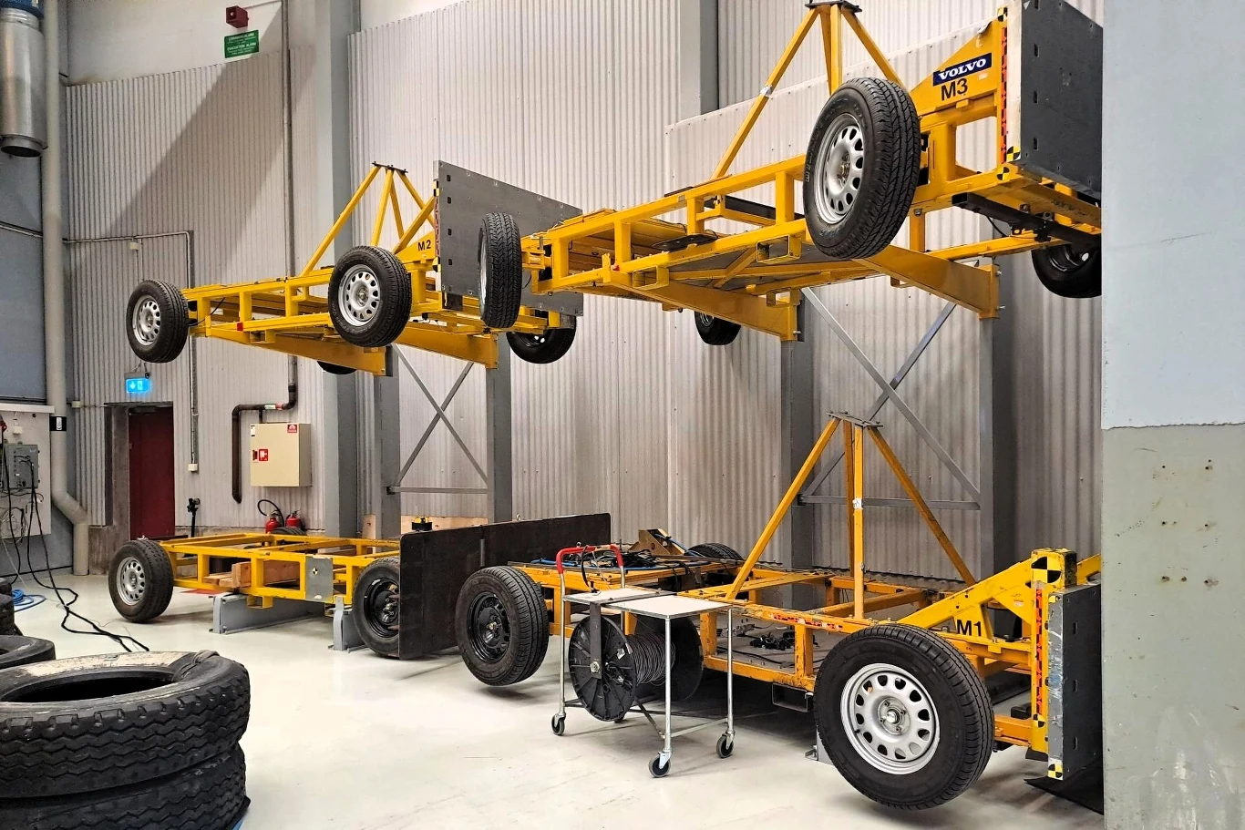 Żółte metalowe ramy wózków widłowych, niektóre zawieszone na dźwigu, inne stojące na posadzce w hali przemysłowej. Widać opony i inne elementy wyposażenia warsztatu lub magazynu. 