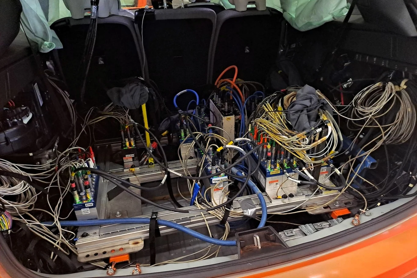 W bagażniku samochodu osobowego znajduje się złożony system elektroniczny. Widoczne są liczne przewody, moduły i urządzenia pomiarowe, co sugeruje, że samochód został przystosowany do celów badawczych lub testowych. 