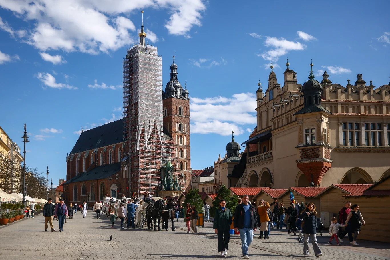 Kościół Mariacki w Krakowie, Sukiennice i plac wokół nich, tętnią życiem. Dorożki czekają na turystów, a ludzie spacerują po brukowanym placu. W tle widać rusztowania na wieży kościoła Mariackiego, co sugeruje prace remontowe lub konserwatorskie. Słone...