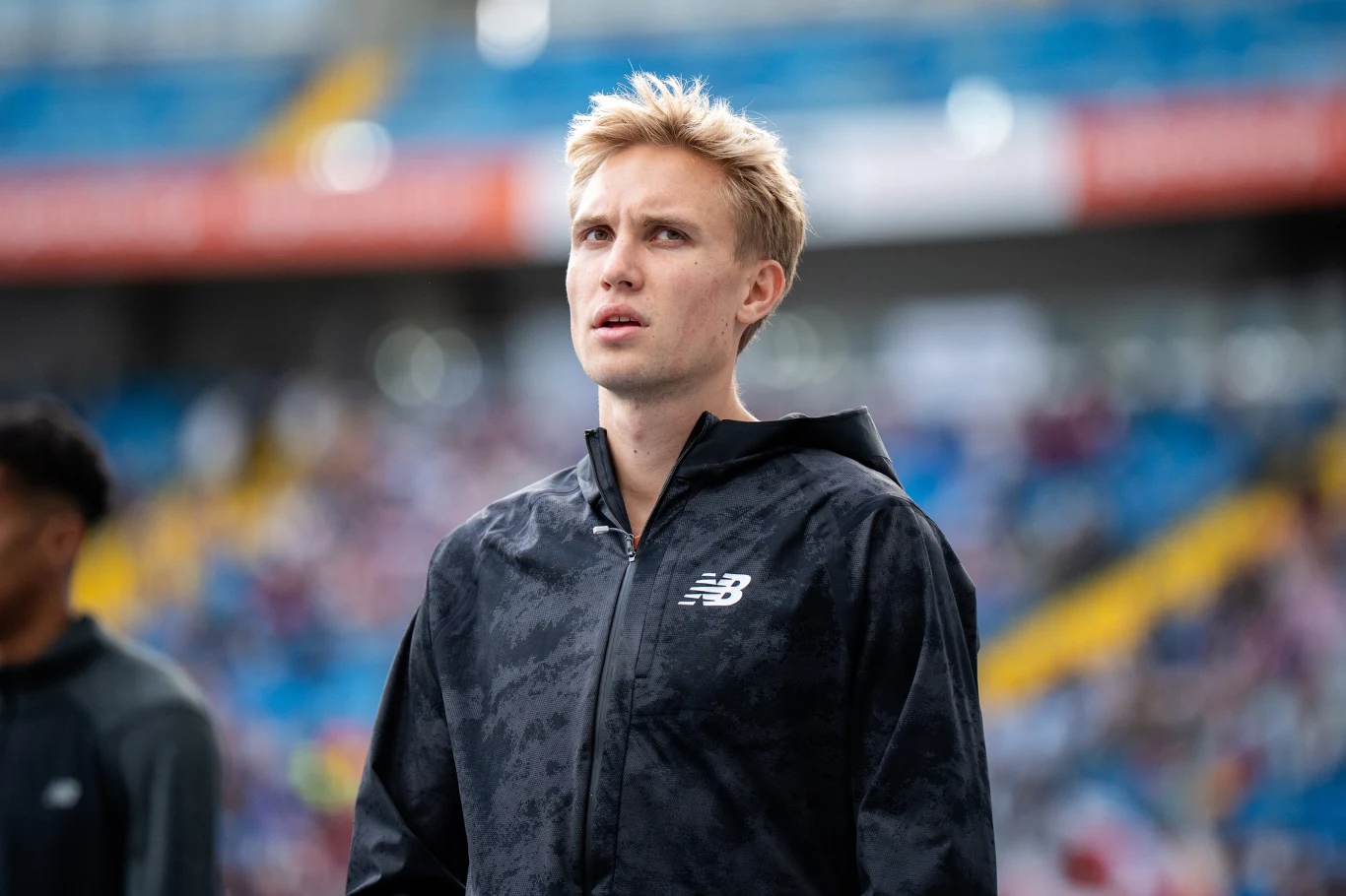 Młody, blondwłosy mężczyzna w czarnej kurtce z logo New Balance na stadionie, spogląda w górę. Na drugim planie widać trybuny stadionu i rozmytą postać innej osoby w podobnej kurtce.