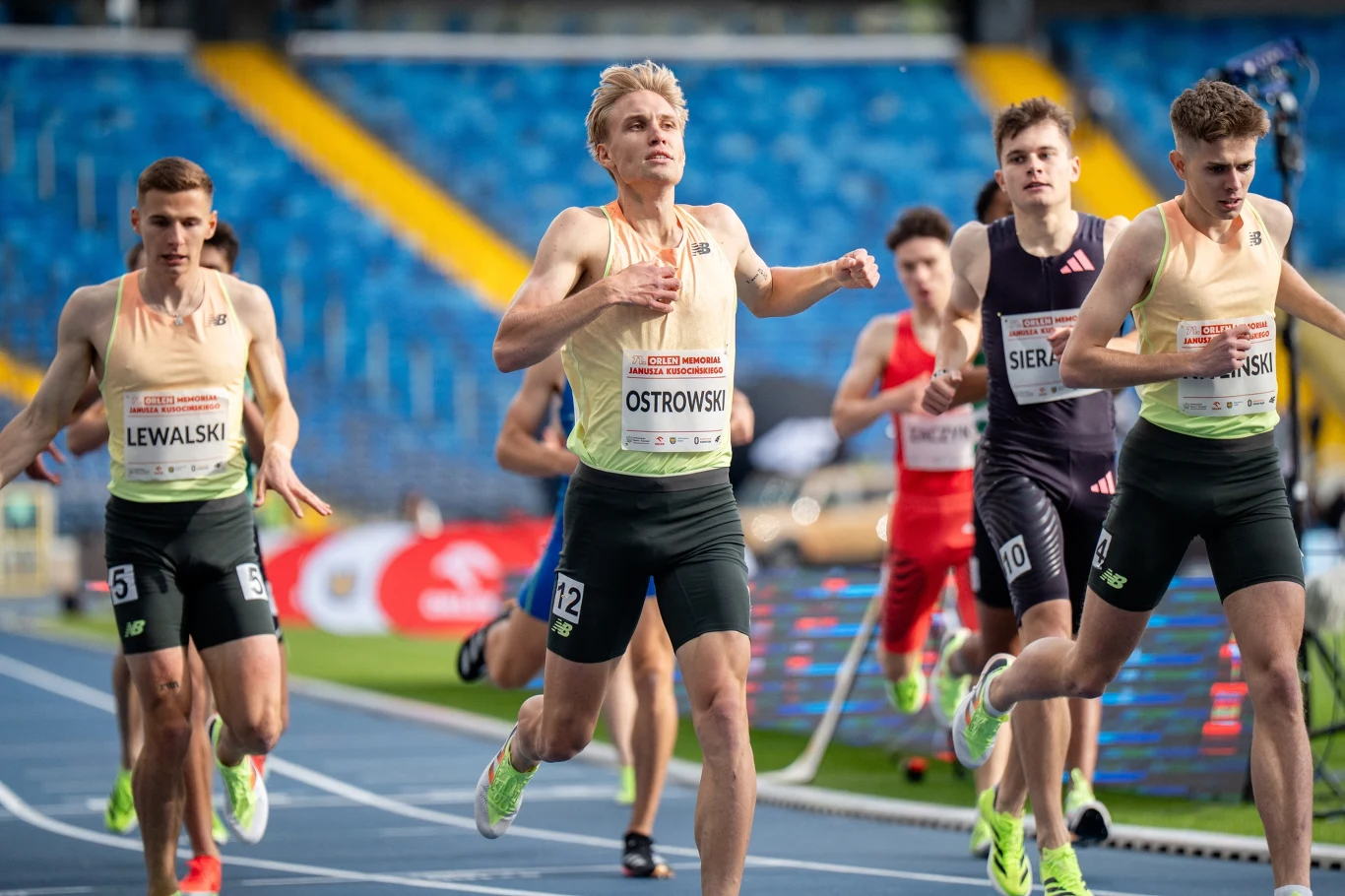 Grupa młodych mężczyzn w sportowych strojach biegnie po bieżni lekkoatletycznej podczas zawodów. Zawodnicy mają na sobie koszulki z nazwiskami i numerami startowymi. Widać skupienie i determinację na ich twarzach, gdy walczą o jak najlepszy wynik. Na p...