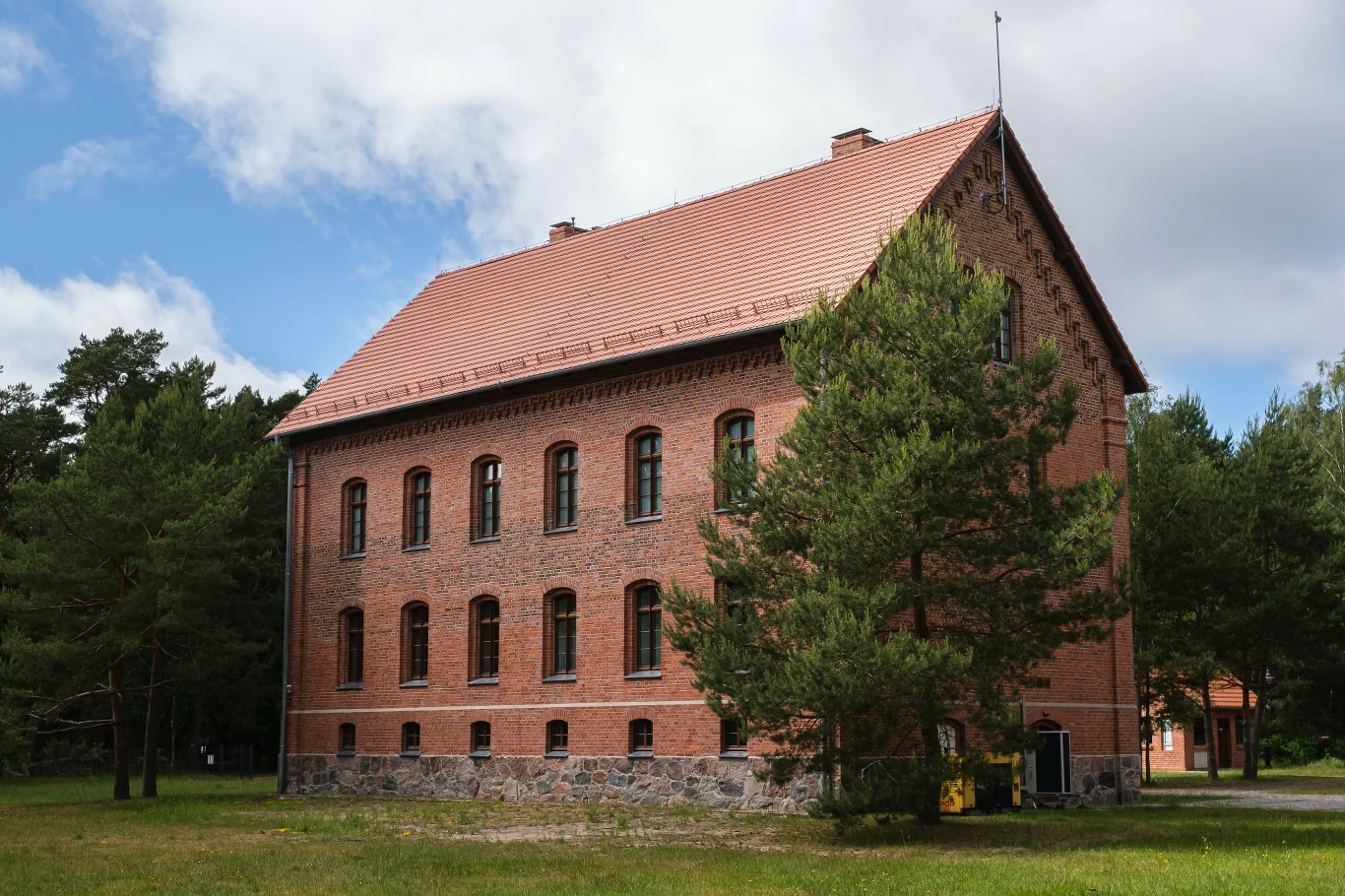Dwupiętrowy budynek z czerwonej cegły z rzędami okien i spadzistym dachem, usytuowany na skraju lasu, widoczny w letni, słoneczny dzień