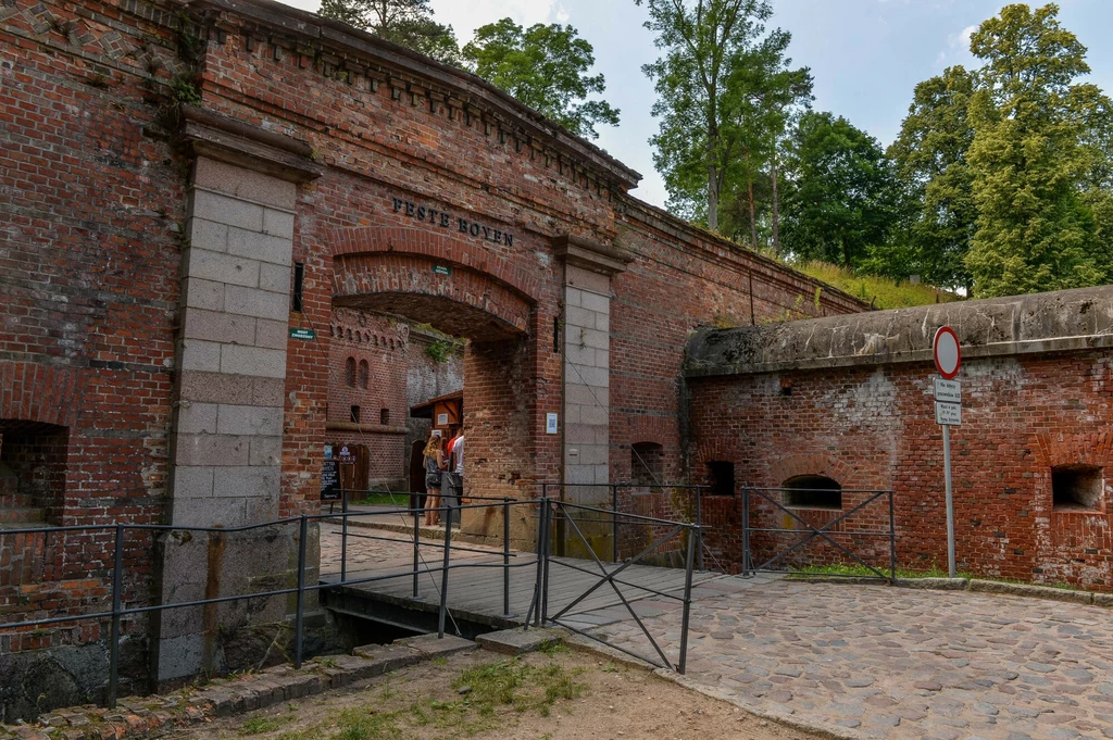 Brama z napisem "Feste Boyen" w Giżycku, Polska. Ceglana brama z białymi, kamiennymi filarami po bokach, prowadzi do wnętrza fortu. Po prawej stronie bramy widoczny jest fragment muru obronnego, a na nim znak zakazu ruchu. Przed bramą znajduje się meta...
