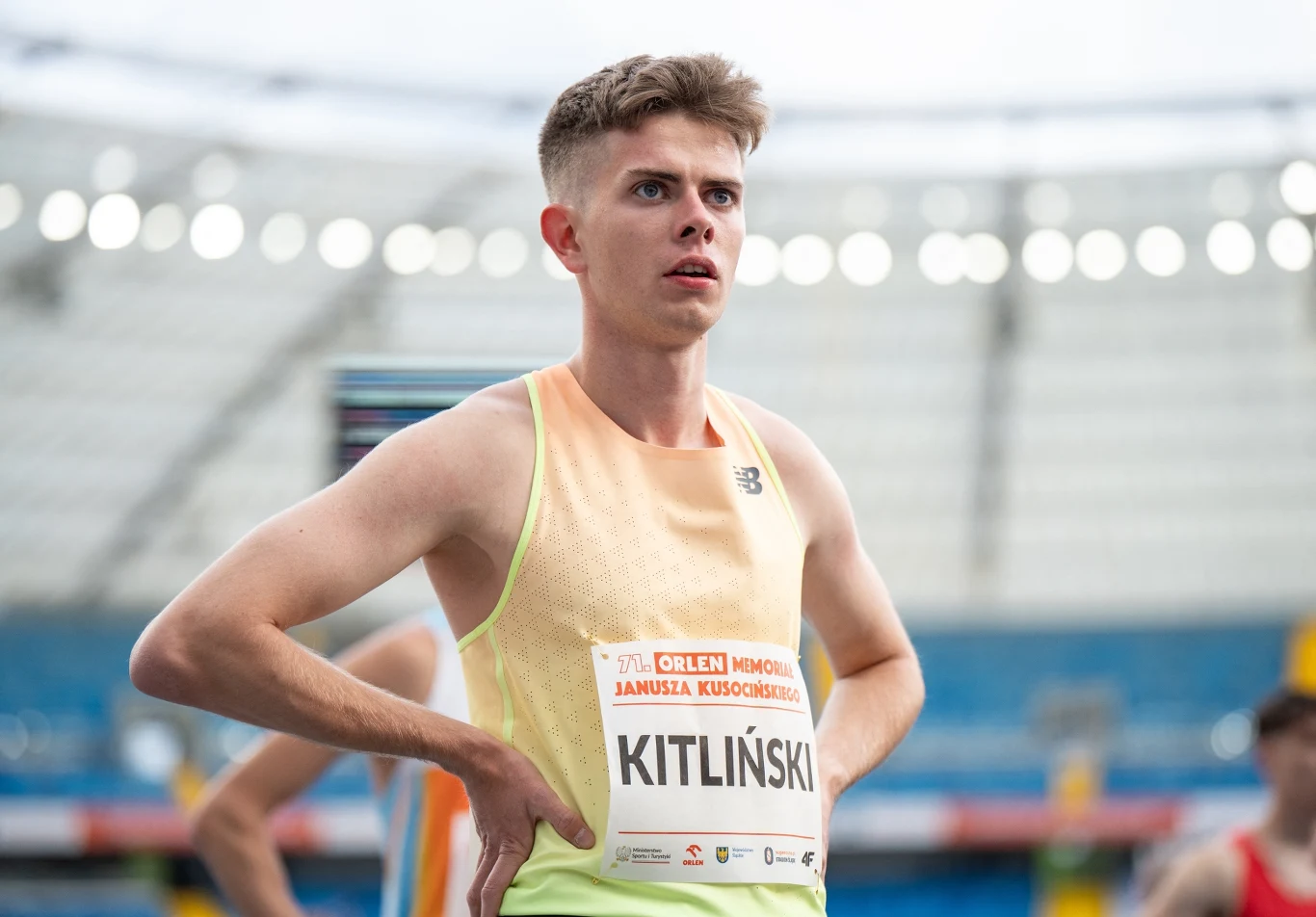 Bartosz Kitliński Zawodnik lekkoatletyczny stoi na bieżni, ręce ma oparte na biodrach, a wzrok skierowany w górę. Ma na sobie żółto-zieloną koszulkę z numerem startowym i nazwiskiem "Kitliński".