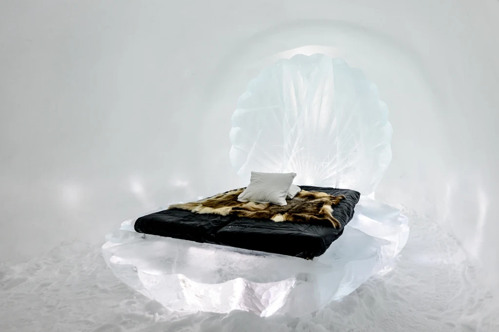 Wyjątkowe hotele na świecie Łóżko w lodowym hotelu, przykryte ciemną kołdrą i futrem, z białą poduszką. Za łóżkiem znajduje się lodowa rzeźba przypominająca muszlę. Podłoga wykonana jest ze śniegu. Pokój hotelowy jest cały z lodu