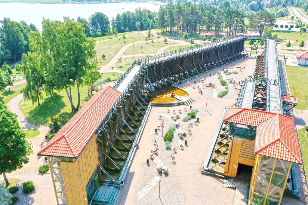 Drewniana konstrukcja tężni solankowej w Augustowie, widziana z lotu ptaka. Tężnia znajduje się w parku, niedaleko jeziora Necko. Wokół tężni rozmieszczone są ławki i leżaki, a także ścieżki spacerowe i roślinność. Zdjęcie ukazuje nowoczesną infrastruk...