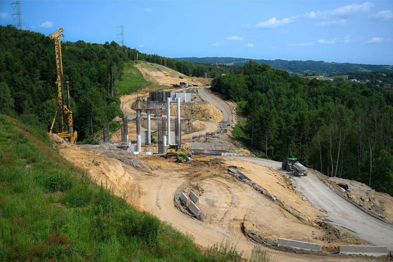 Droga ekspresowa S19 będzie częścią trasy Via Carpatia. Budowa drogi lub wiaduktu w terenie pagórkowatym. Widoczne są filary, maszyny budowlane i ciężarówka. Część terenu jest zalesiona, a część pokryta ziemią i roślinnością. W tle widać linię energetyczną i dalszy krajobraz