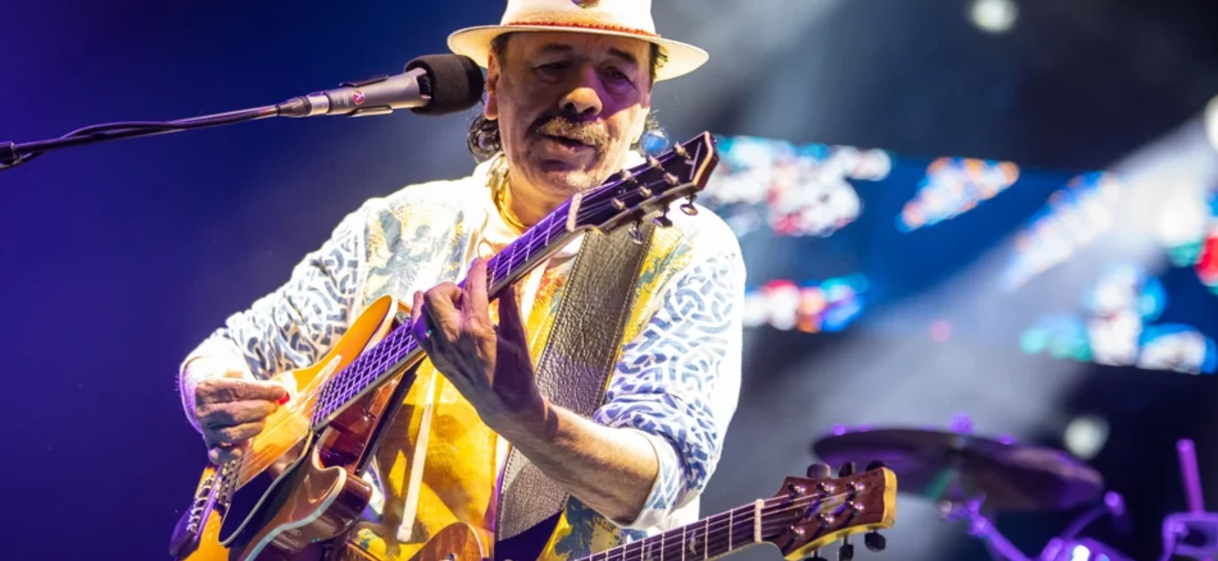 Koncert Santany w Łodzi Gitarzysta w kapeluszu gra na gitarze na scenie, oświetlony reflektorami. Na drugim planie widać perkusję i rozmyty tłum ludzi w kolorowych światełkach scenicznych. Muzyk skupiony jest na grze, a jego ręce poruszają się po gryfie gitary. Na scenie panu...
