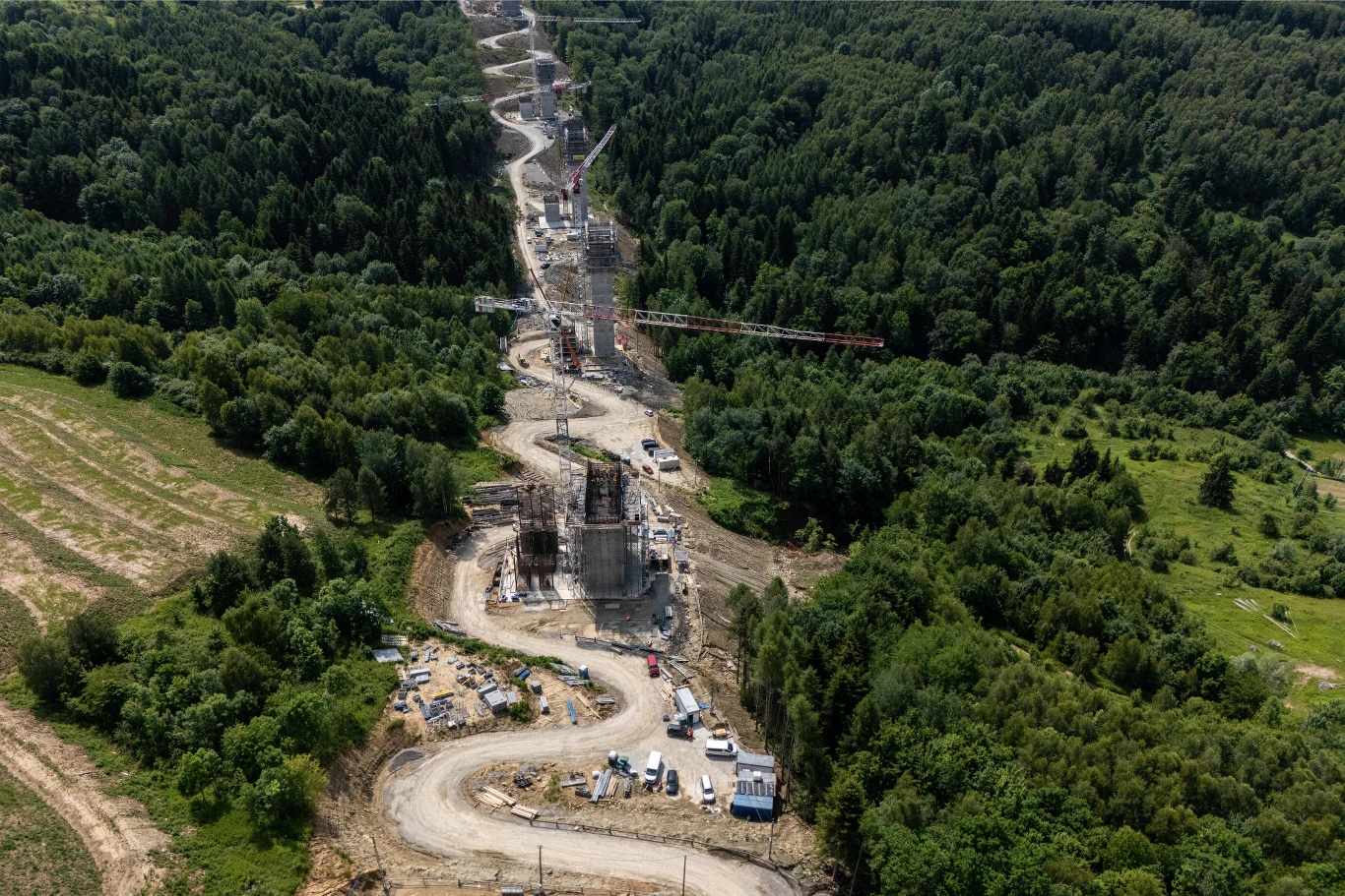 Odcinek Babica - Jawornik jest wyjątkowo wymagający. Powstanie osiem estakad, pięć wiaduktów i cztery przejścia dla zwierząt. Budowa wiaduktu drogowego w lesie. Widoczne dźwigi, pojazdy budowlane i betonowe filary. Droga wije się przez teren budowy, otoczona zielenią drzew. Zdjęcie wykonane z lotu ptaka ukazuje skalę przedsięwzięcia i postęp prac. Na placu budowy znajdują się...