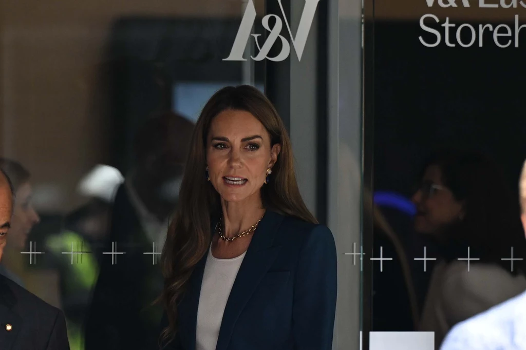 Odwiedziny Kate nieco zaskoczyły fanów Kate Middleton, księżna Walii, uśmiecha się lekko, stojąc przed wejściem do budynku. Ma na sobie granatową marynarkę i biały top, a jej włosy są rozpuszczone. Na szyi ma złoty naszyjnik. W tle widać fragment logo sklepu & Other Stories, z refleksami na...
