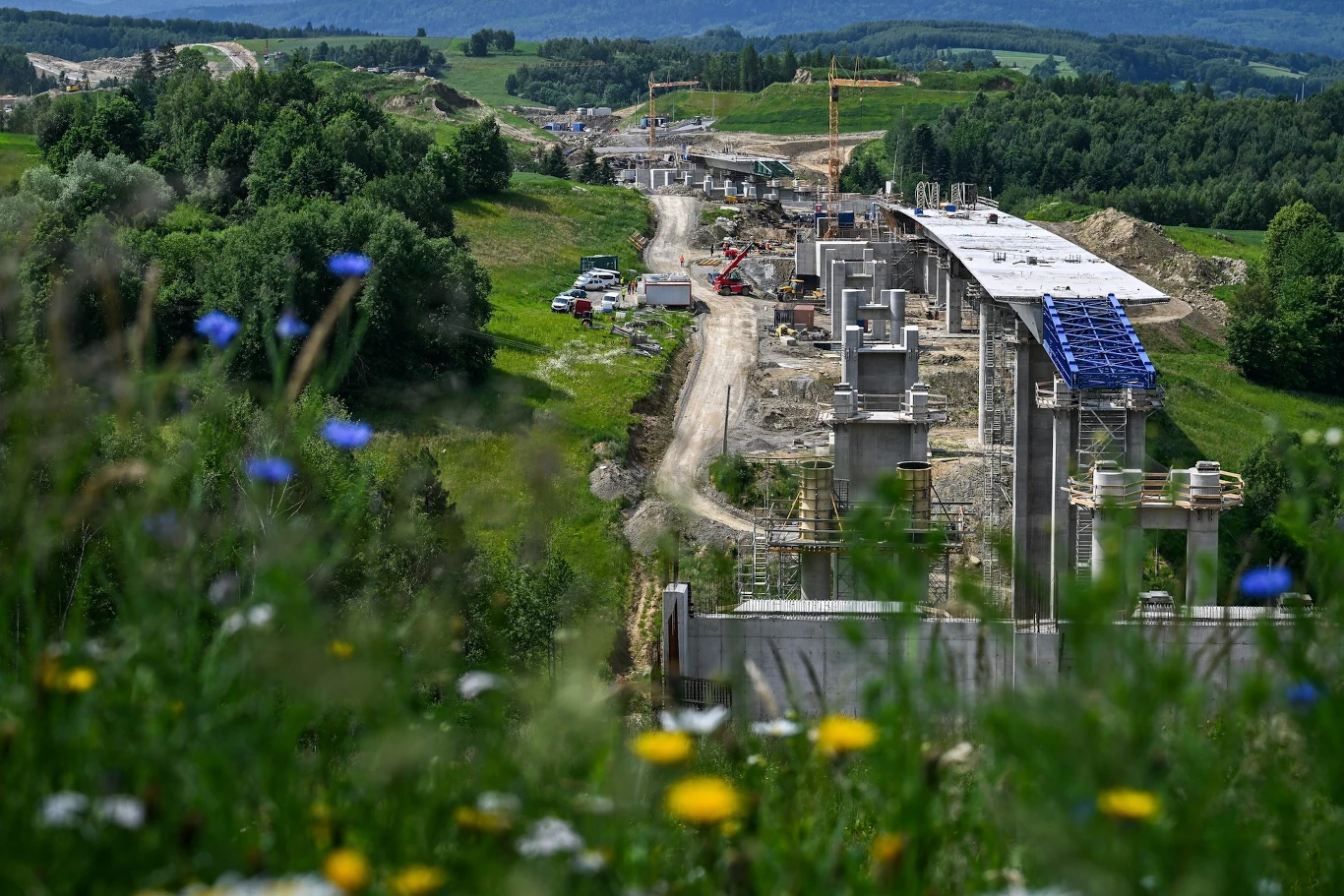 Budowa drogi S19 Rozległy plac budowy wiaduktu lub mostu prowadzącego przez pagórkowaty teren, w otoczeniu zielonych lasów i łąk z widocznymi maszynami budowlanymi oraz betonowymi konstrukcjami.