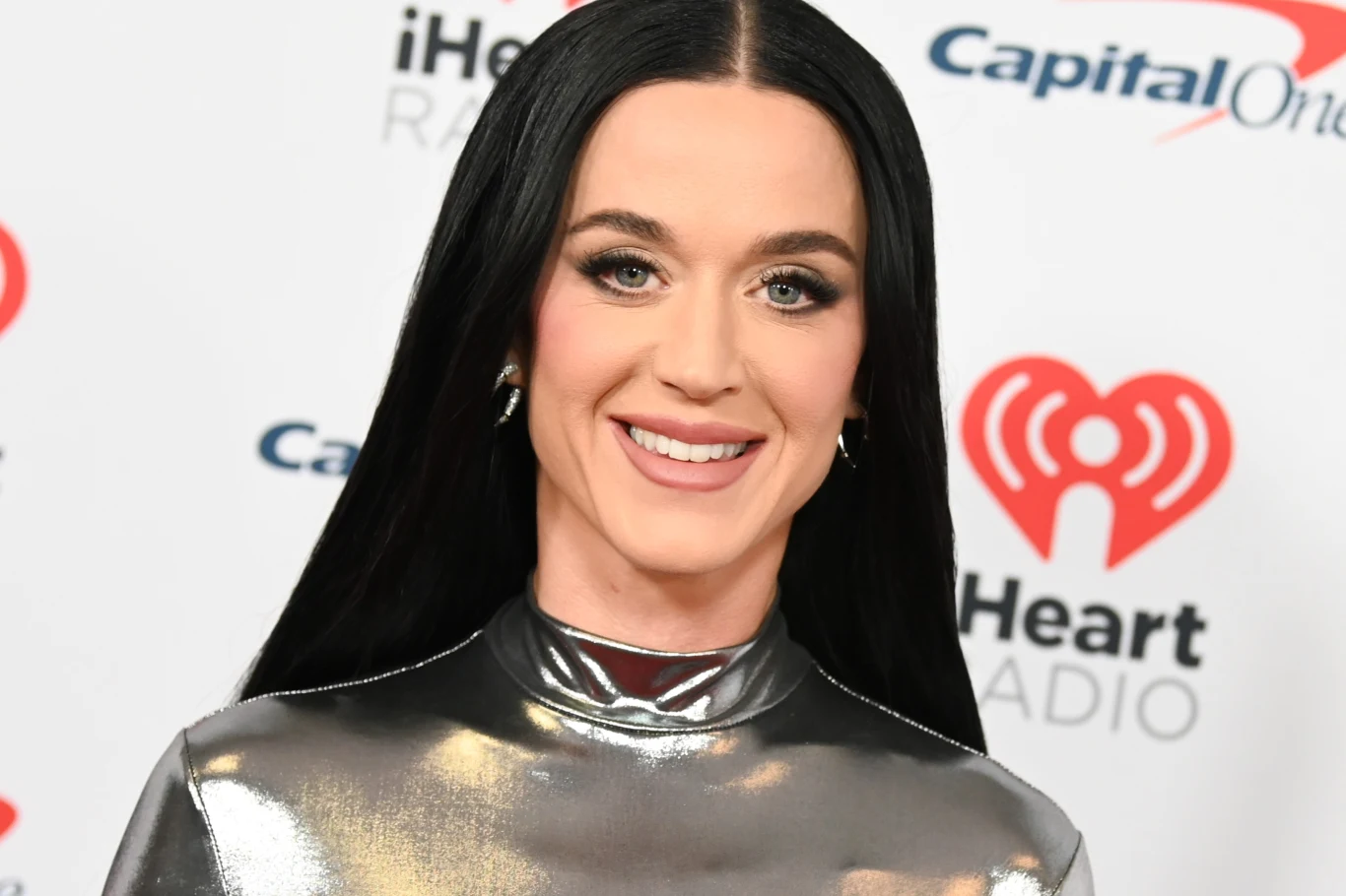 Katy Perry wystąpiła ostatnio w Australii Kobieta z długimi, czarnymi włosami uśmiechająca się w metalicznej, srebrnej bluzce, stojąca na tle ściany z logotypami iHeart Radio oraz Capital One.